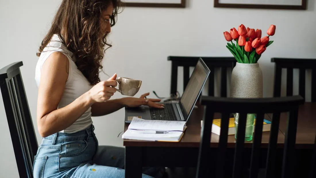 A woman sitting at a dining table typing on a laptop with a cup of coffee in her other hand, a vase of red tulips beside her.