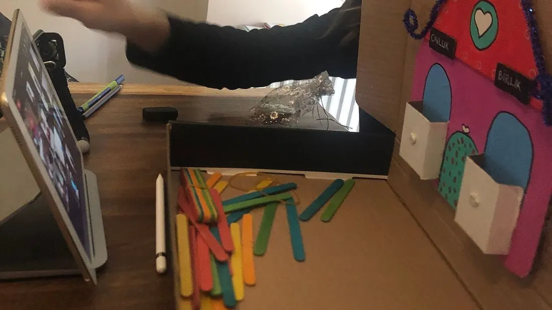 A teacher seen from the side at a wooden desk with a tablet showing a video lesson on the left and a handmade cardboard house with coloured sticks in front of it.
