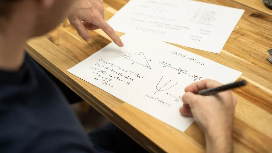 Two people at a wooden table with worksheets of mathematics and chemistry; one hand points at a worksheet while the other holds a pen.