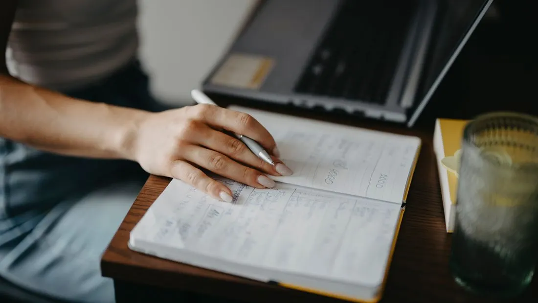 A hand holding a white pen over an open notebook full of handwritten notes, with a laptop and a glass of water on the wooden desk.