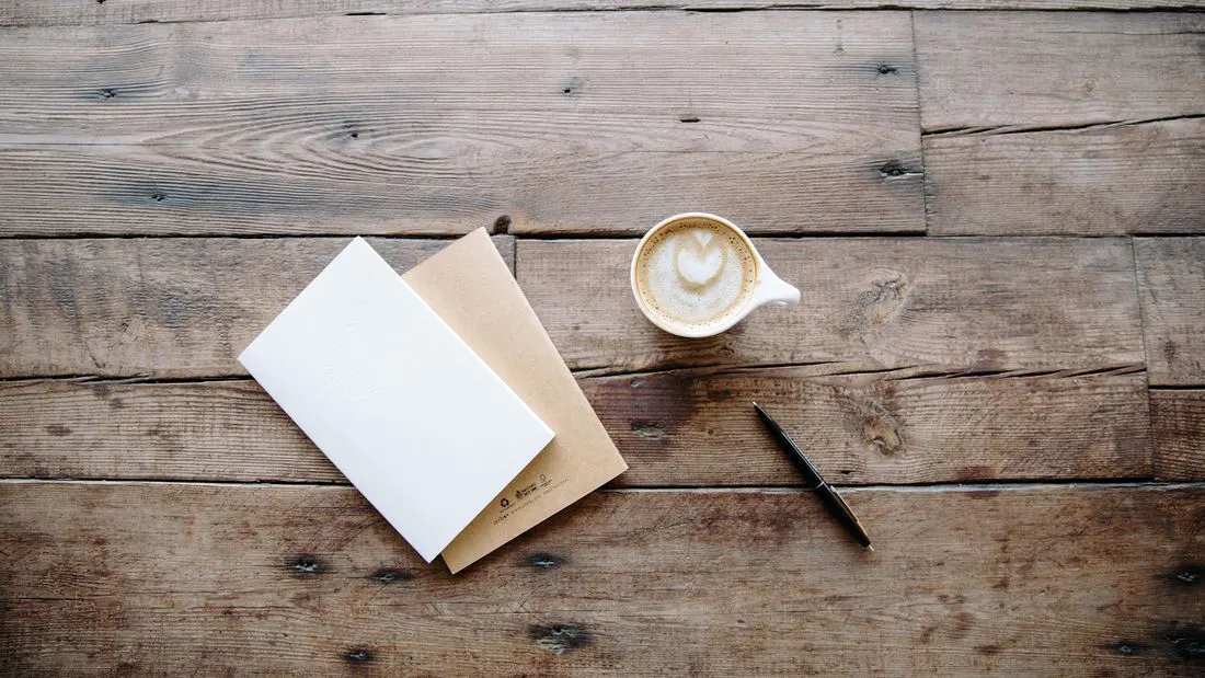 A wooden table seen from above with a cup of coffee, two closed notebooks and a pen.