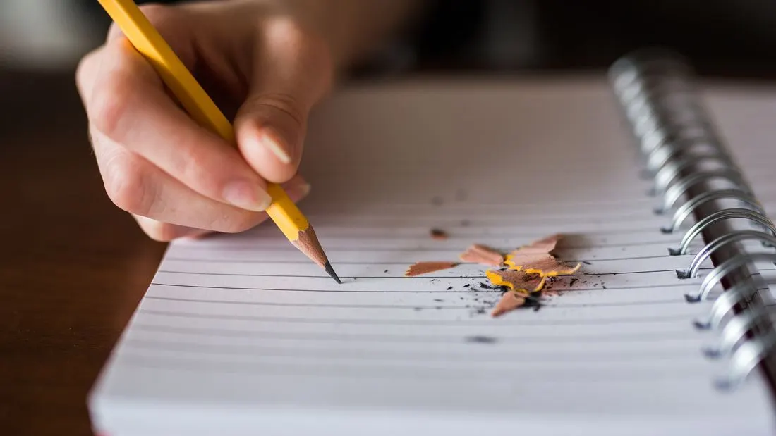 A hand holding a pencil over a notebook page of handwritten notes on a table.