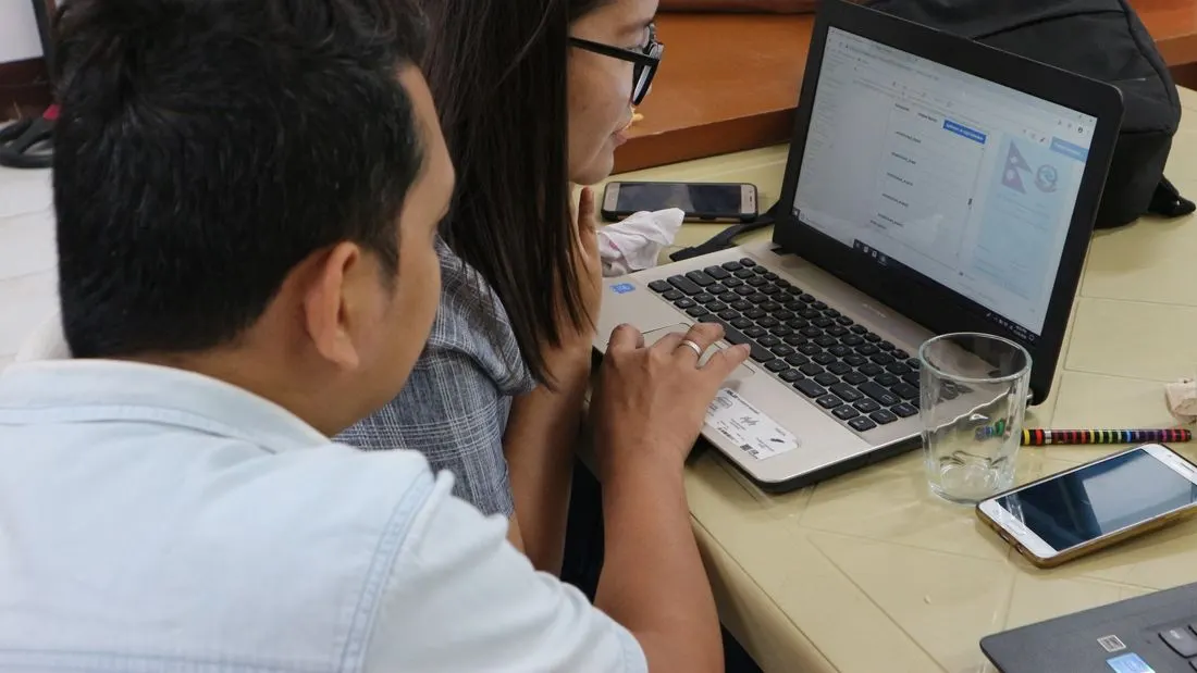 Two people seen from behind looking at a laptop screen on a table, one of them touching the trackpad, with a glass, a pencil and two phones on the table.