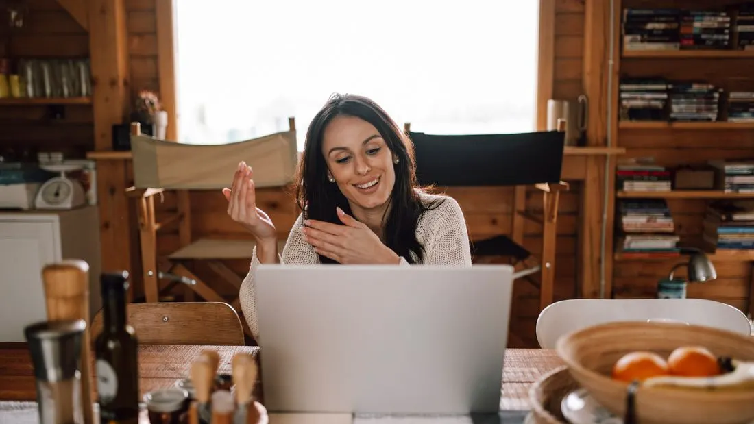 A smiling woman with long dark hair talking and gesturing toward an open laptop on a wooden table in a bright room with bookshelves behind her.
