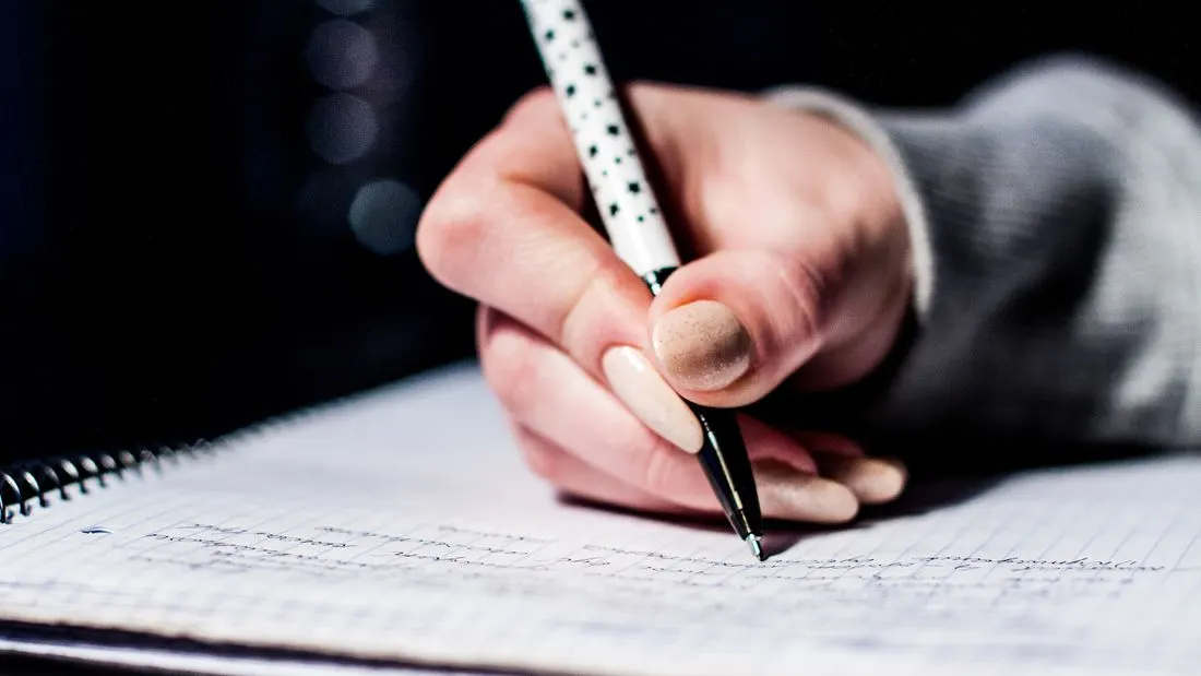 A hand holding a white pen with black stars, writing notes in a spiral notebook on a dark background.