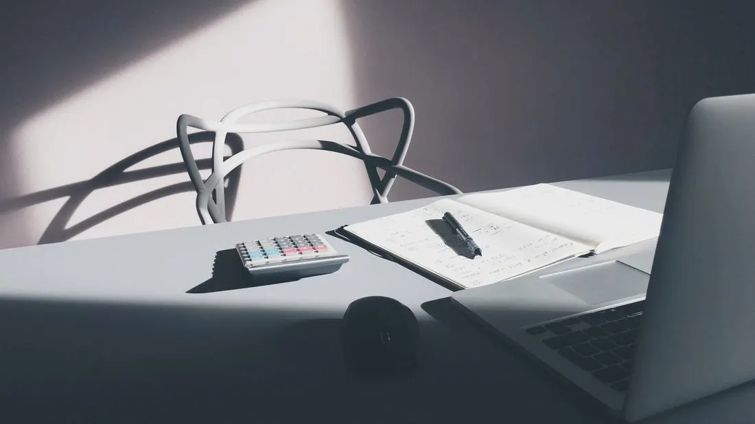 A desk in a patch of sunlight with a small calculator, an open notebook with a pen and the edge of a laptop.