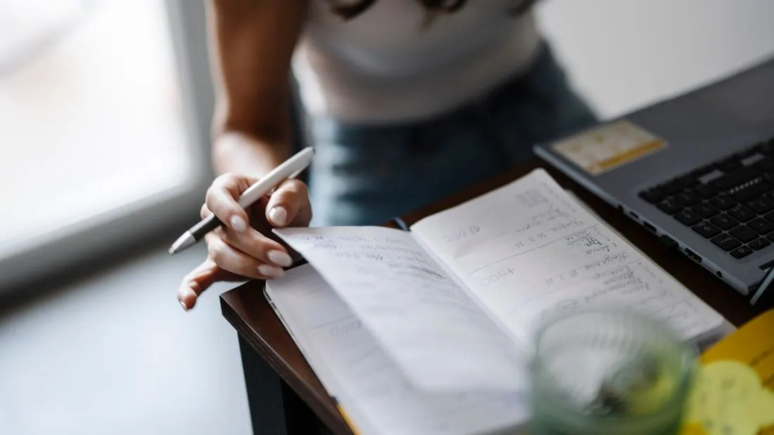 A person at a desk with a laptop open in front of them holding a pen over a notebook full of handwritten notes, with a glass of water beside them.