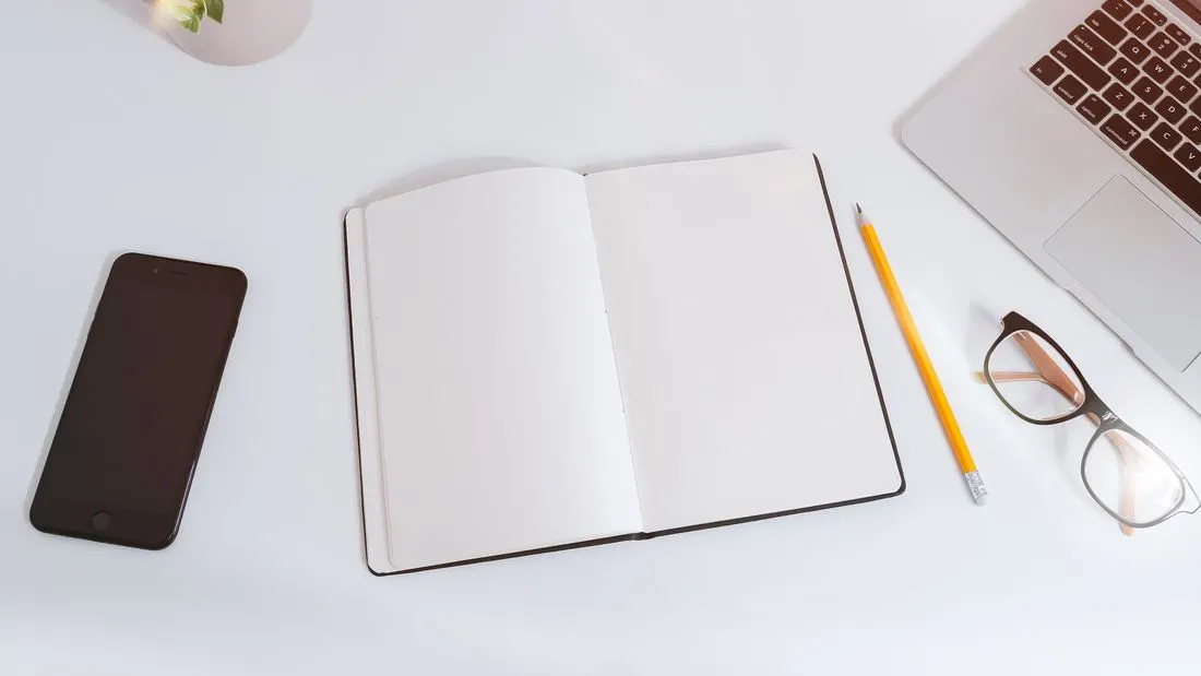 A white desk seen from above with an open blank notebook, a pencil, a pair of glasses, a smartphone and the corner of a laptop.