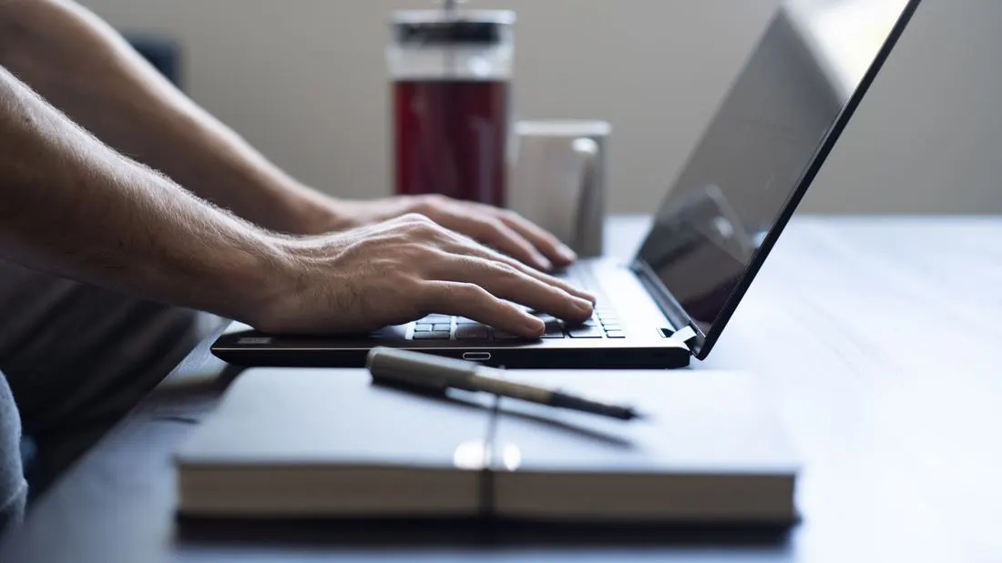 Close view of a person typing on a black laptop beside a closed notebook with a pen and a glass jug of dark red tea.
