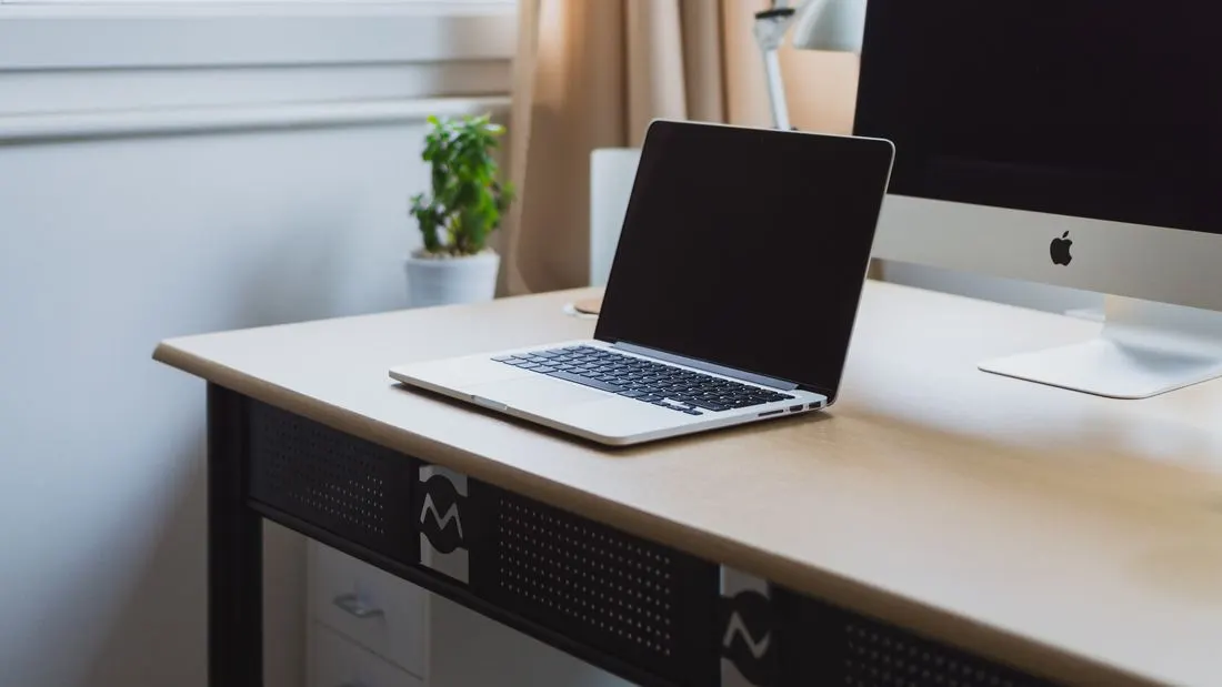 A silver laptop on a pale wooden desk beside a potted plant, with a desktop computer screen at the edge of the picture.