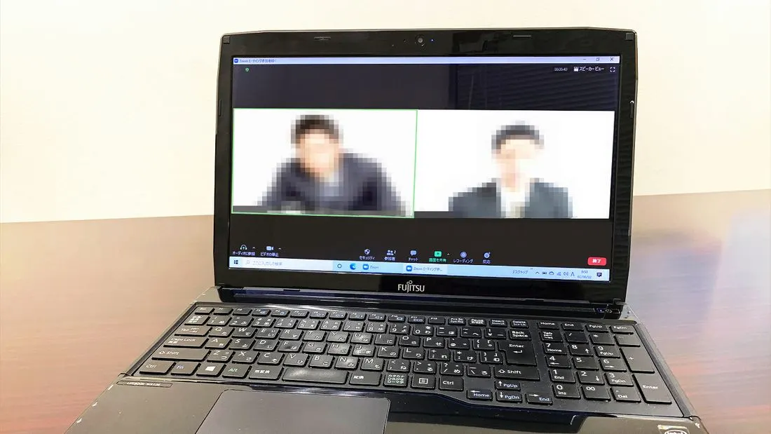 An open laptop on a dark wooden table showing a video call with two people, their faces blurred.