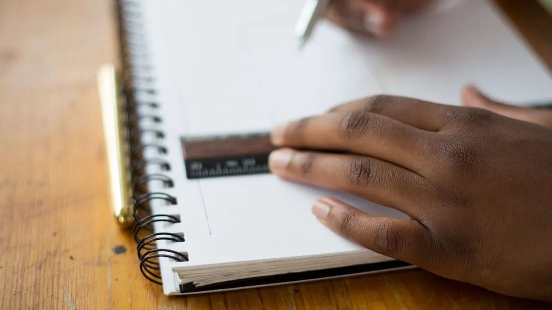 A hand holding a short ruler flat against a blank page of a spiral notebook, with a pen held above it.