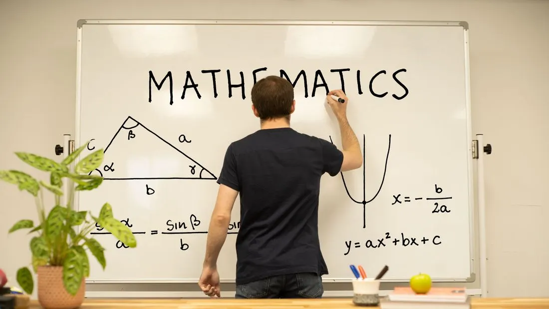 A person seen from behind writing the word MATHEMATICS on a whiteboard with a labelled triangle, a parabola and the quadratic formula drawn beside it.