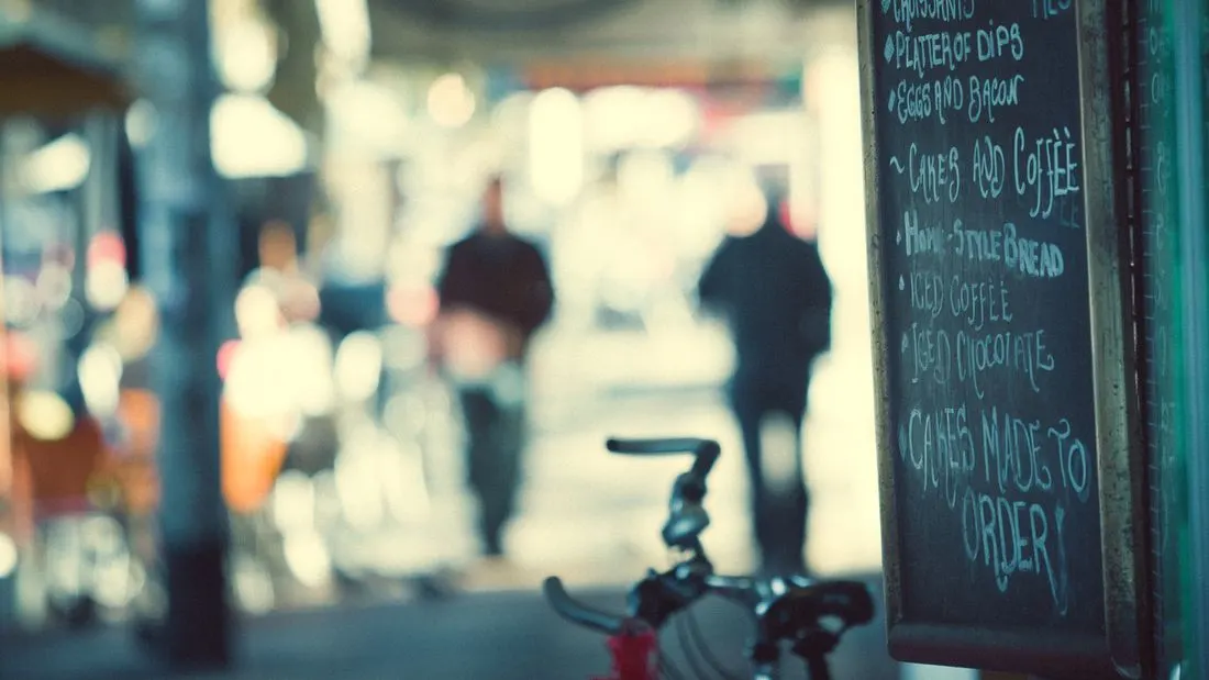 A small chalkboard sign with handwritten menu lines stands on the right of the frame, with blurred walkers and a bicycle handlebar behind it.