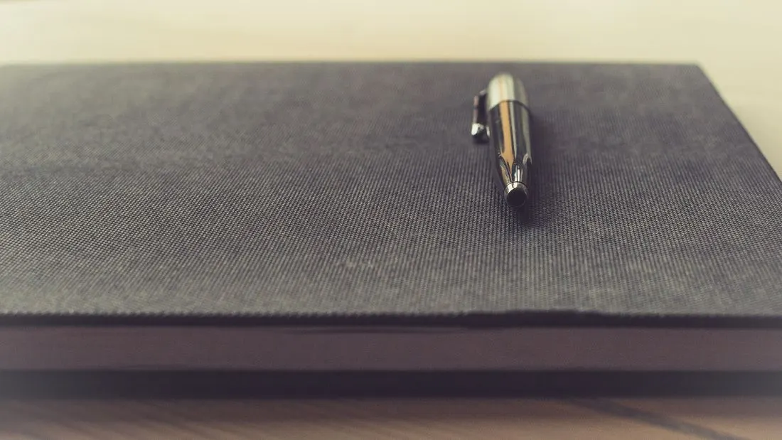 A closed dark cloth-bound notebook on a desk with a silver pen lying across the cover.
