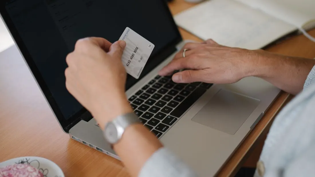 A person at a desk holding a plastic card in one hand and typing on a laptop with the other.