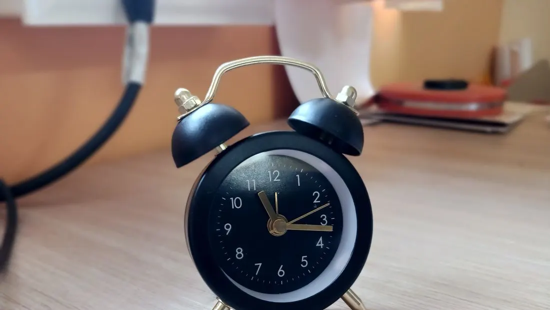 A small black alarm clock with gold hands standing on a light wooden desk.
