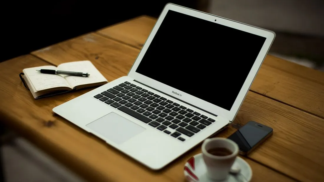 A laptop, an open notebook with a pen, a phone and a cup of coffee on a wooden table.
