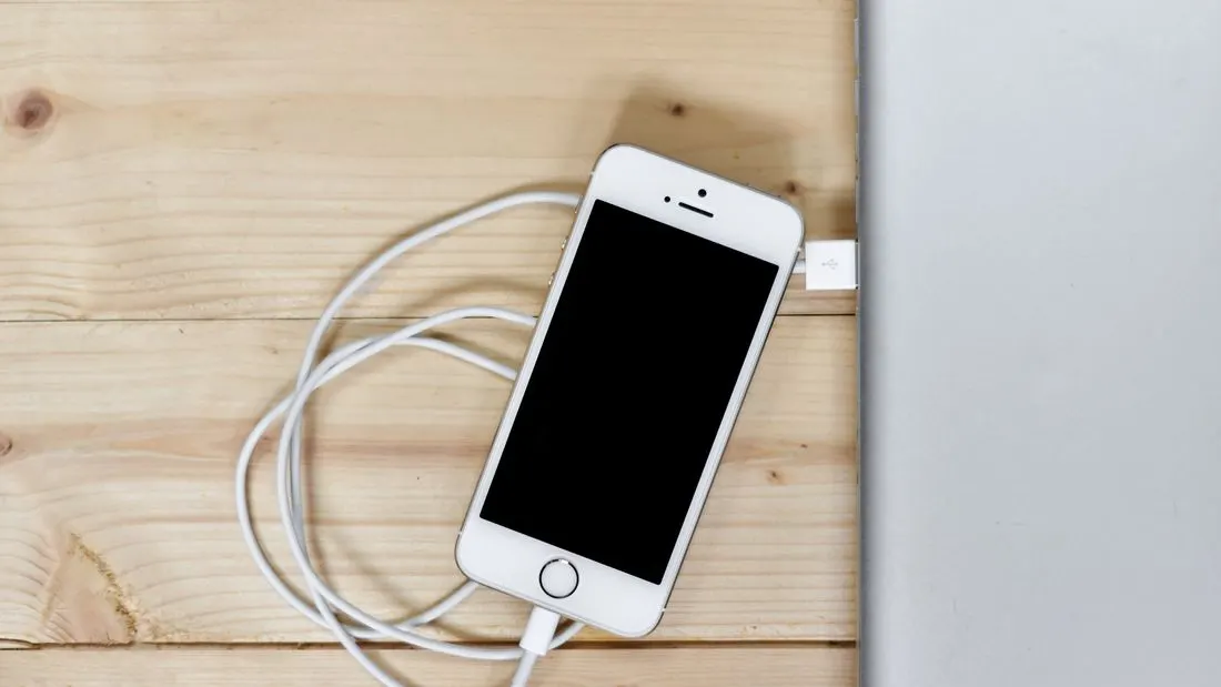 A smartphone lying on a pale wooden desk beside a laptop, connected by a white cable.