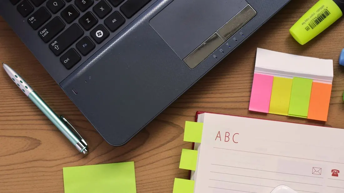 A laptop, a pen, sticky notes and a planner with coloured tabs on a wooden desk, a phone in the corner.