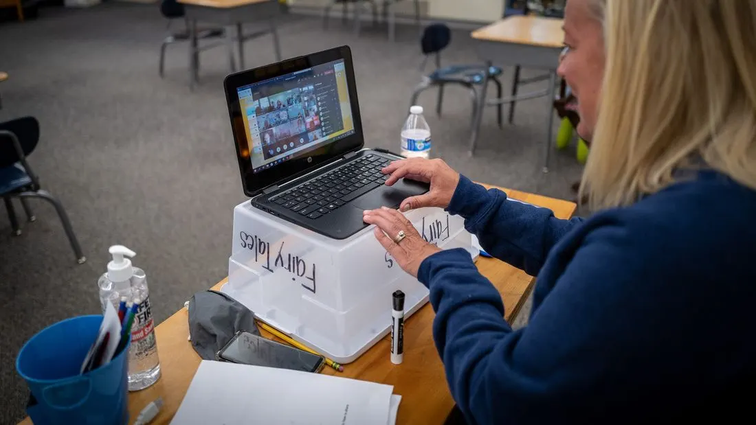 A teacher at a desk using a laptop whose screen shows a video lesson with many small faces.