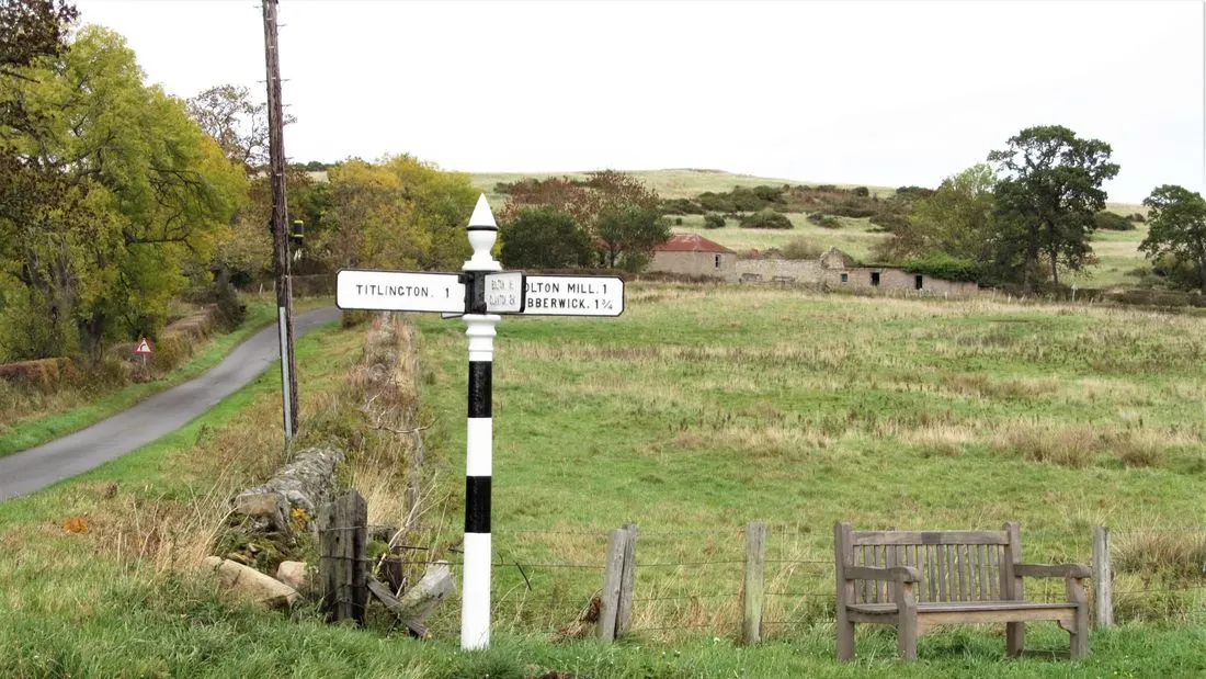 A black and white signpost with two white arms stands at a country crossroads beside a wooden bench and a field.