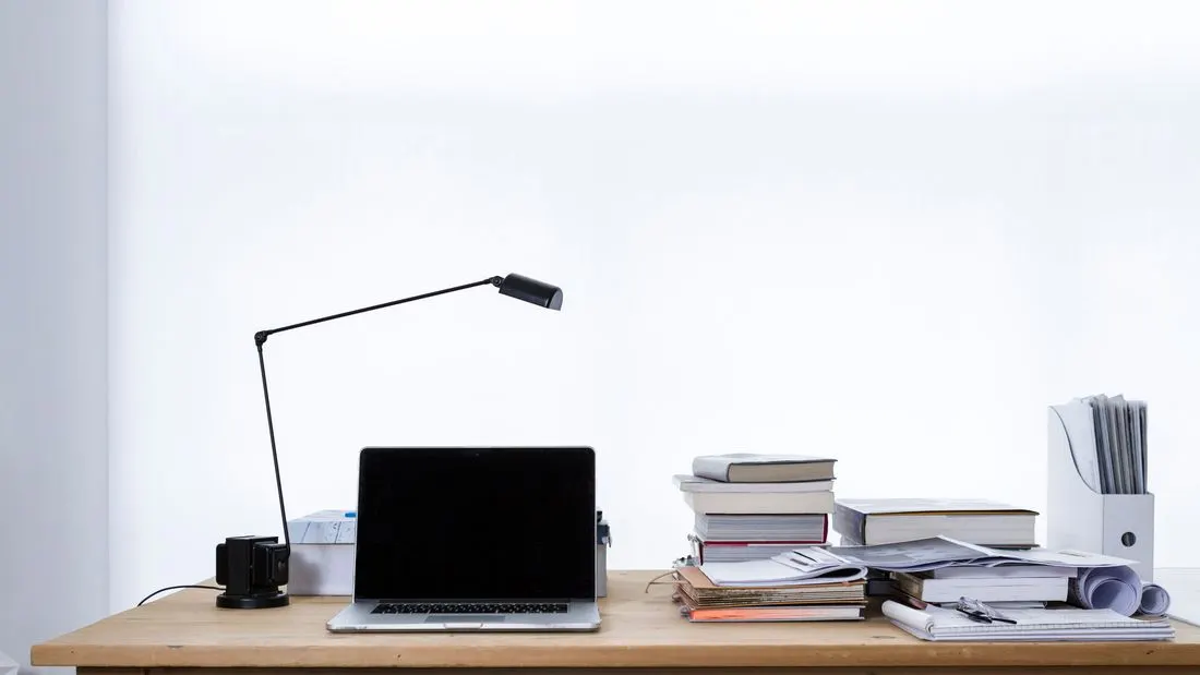 A laptop with a dark screen on a wooden desk beside a black desk lamp and stacks of books, papers and a document holder against a white wall.