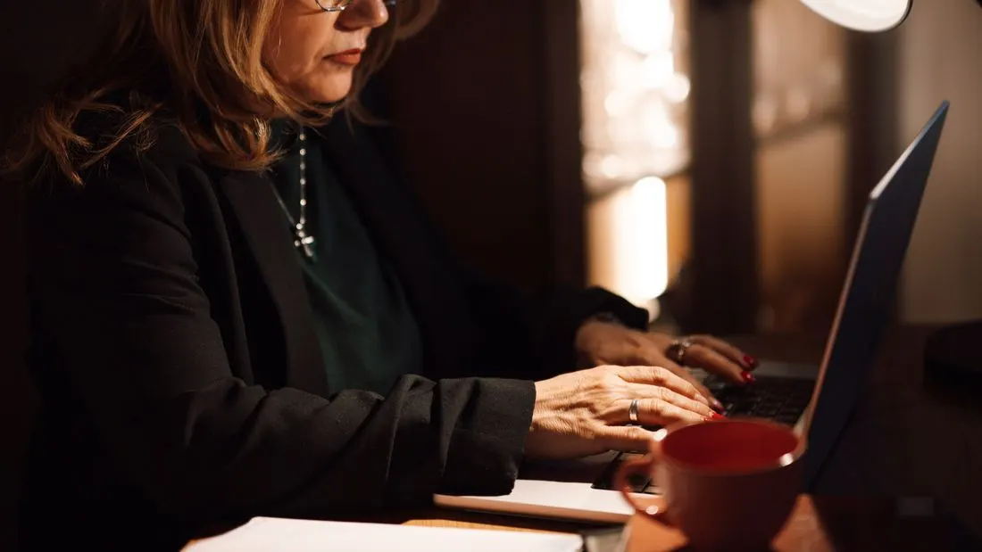 A woman with glasses in a dark jacket types on a laptop at a desk under a lamp, with a red mug beside her.