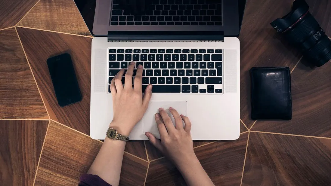 Two hands on a laptop keyboard seen from above, with a phone, a wallet and a camera lens on a wooden desk.
