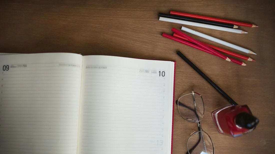 An open diary with blank lined pages on a wooden desk, with colored pencils and a pair of glasses beside it.