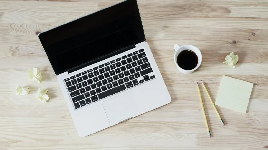 A laptop with a dark screen, a cup of black coffee, two pencils, a pad of paper and crumpled paper balls on a light wooden desk, seen from above.