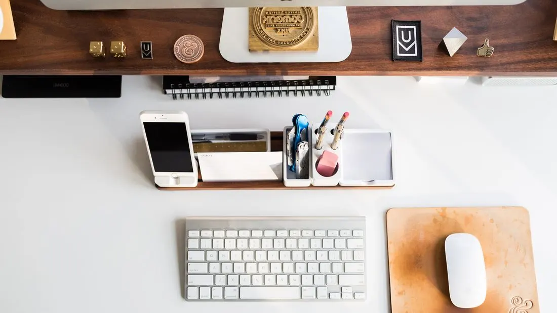A tidy white desk seen from above with a keyboard, a mouse on a leather pad, a phone stand, a spiral notebook and a tray of pens.
