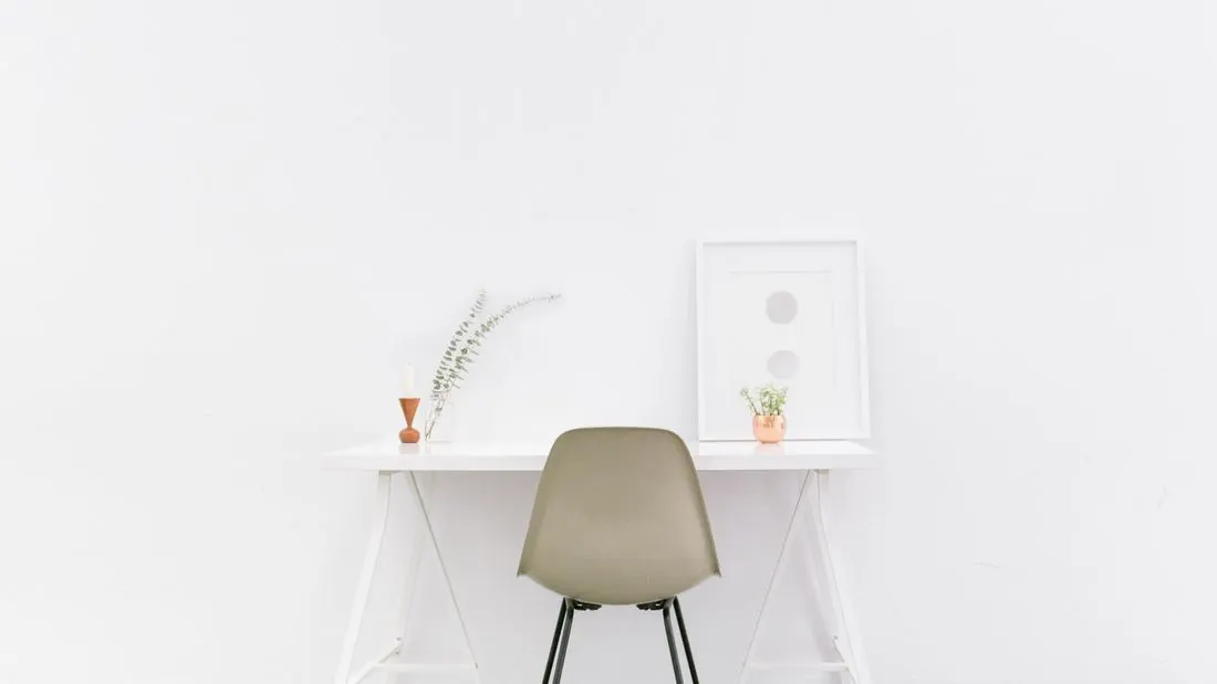 A white trestle desk against a white wall with a plain olive chair, a framed print, a small plant and a candle.