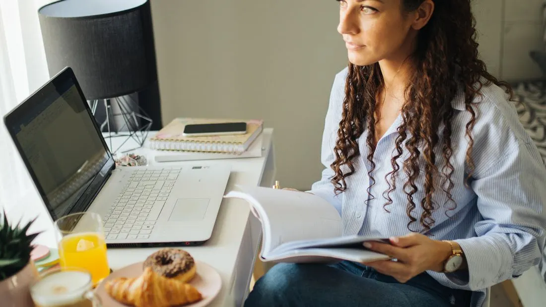 A woman with long curly dark hair sits at a white desk with an open laptop, a notebook, a phone, a croissant and a glass of orange juice, holding an open book and looking away.