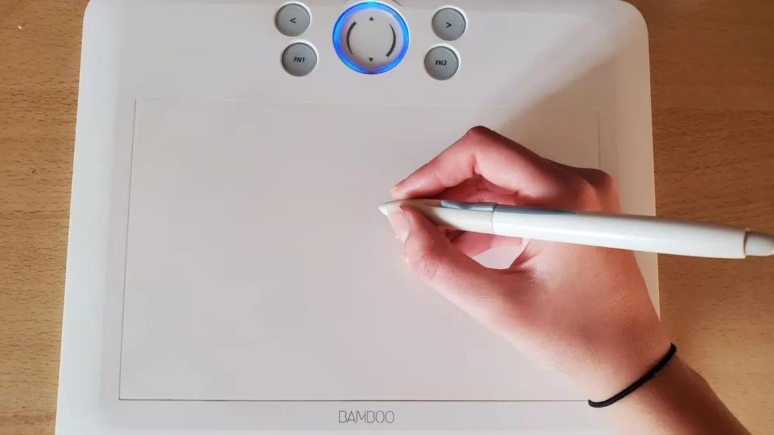 A hand holding a pen over a white graphic tablet on a wooden desk, with a cable and four buttons above the drawing area.