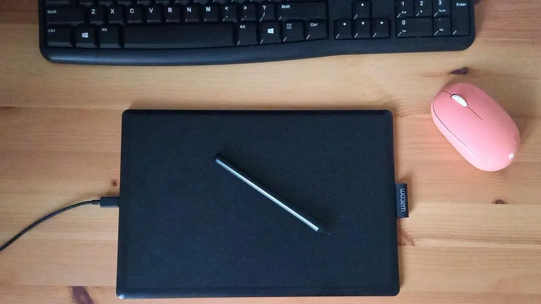 A black pen tablet with a pen on it, a keyboard and a pink mouse on a wooden desk, seen from above.
