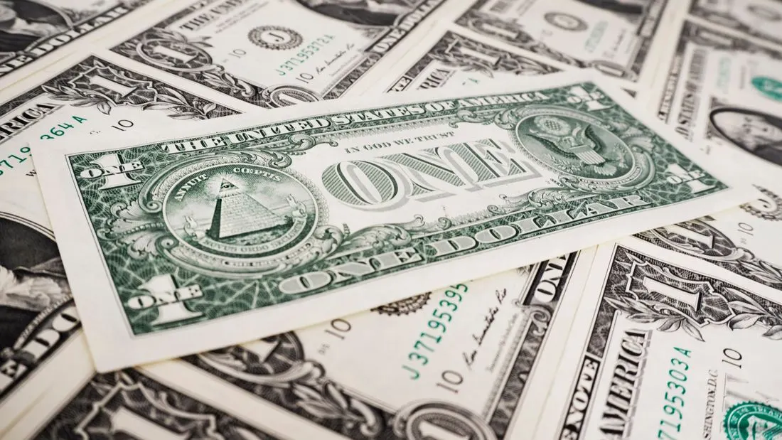 A one dollar bill lying on a pile of other one dollar bills, with the words In God We Trust and One Dollar visible on the back of the top note.