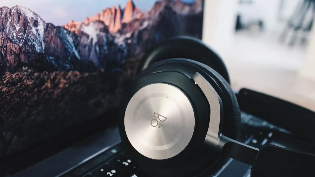 A pair of black over-ear headphones with silver ear cups resting on the keyboard of an open laptop, with a mountain landscape on the screen.