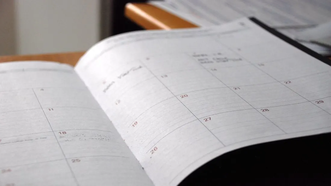 An open paper monthly planner lying on a wooden desk, with a grid of days and a few handwritten notes.